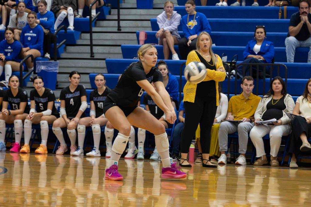 A female volleyball player in a black jersey prepares to receive the ball on the court, with teammates and spectators in the background. A coach stands nearby, observing the play.