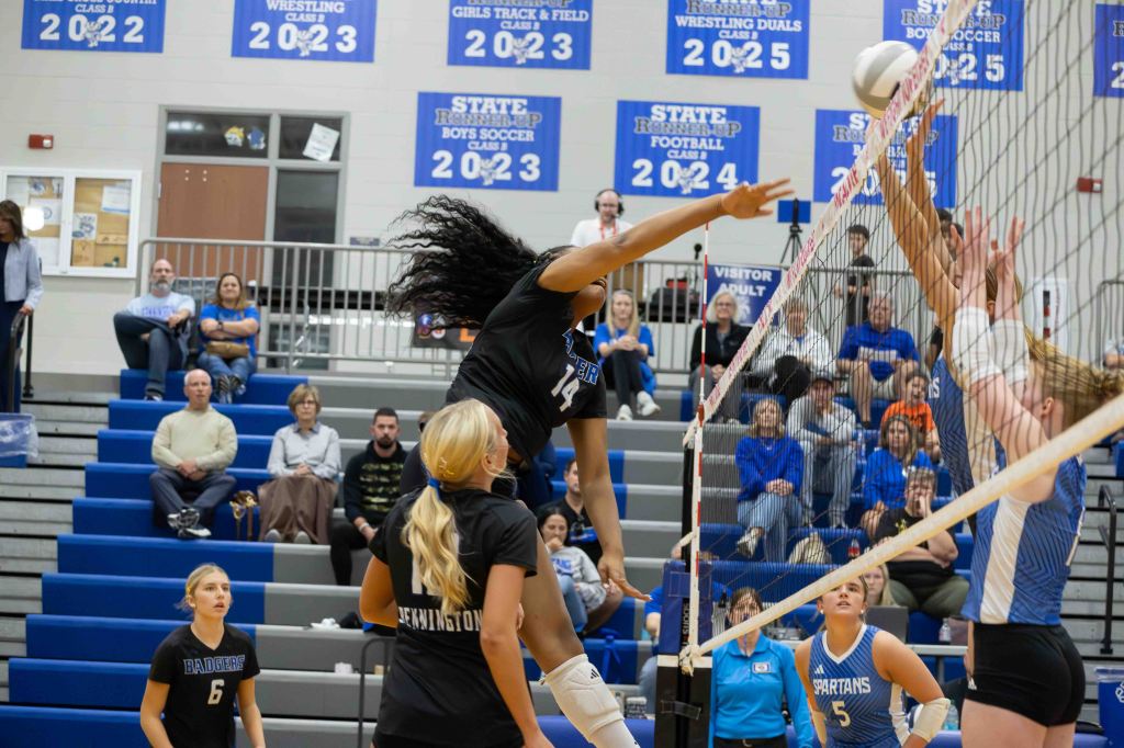A volleyball player leaps to spike the ball over the net during a match, with teammates and opponents nearby. Spectators are visible in the background.