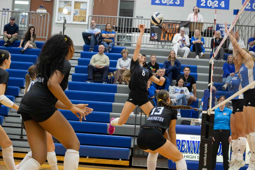 A volleyball player in a black jersey jumps to hit the ball over the net while other players and spectators watch in the background.