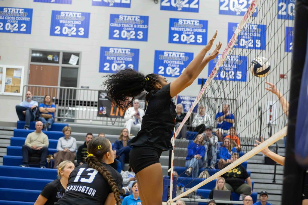 A volleyball player jumps to block a ball at the net, with spectators in the background and team members on the court.