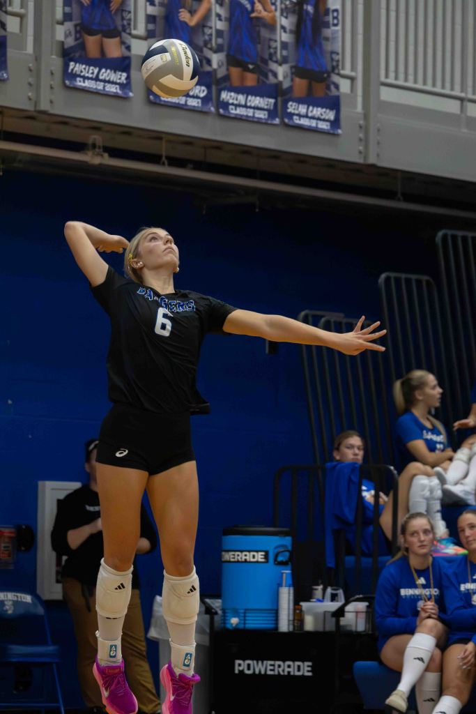 A female volleyball player wearing a black jersey with the number 6 is jumping to serve a volleyball in a gym. In the background, there are banners of teammates on the wall and other players sitting on the bench.