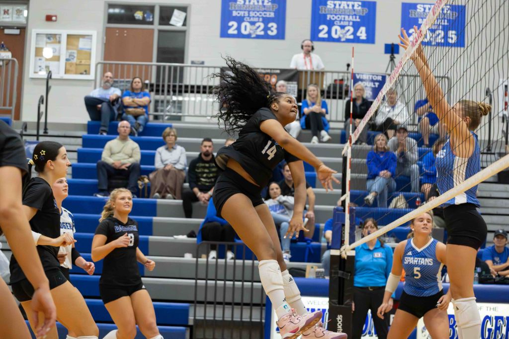 A dynamic volleyball match in progress, featuring a player in a black jersey jumping to spike the ball over the net, while another player in a blue jersey prepares to block. Spectators are visible in the background, watching intently.