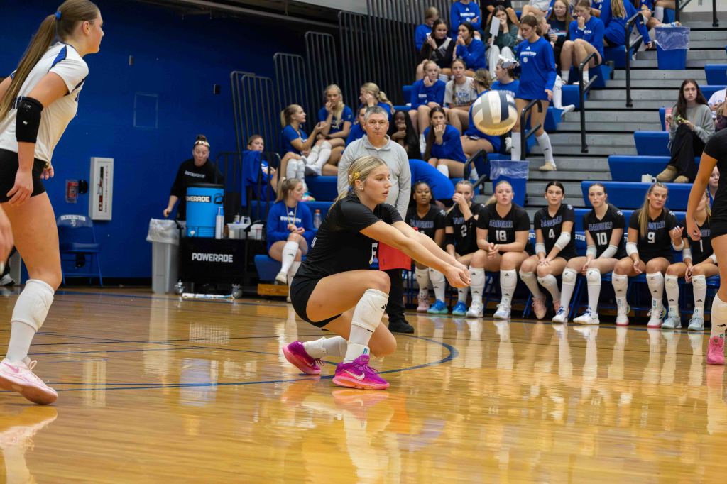 A volleyball player crouches to receive the ball during a match, with teammates and spectators in the background.