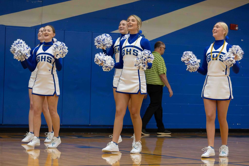 A group of cheerleaders performing energetically while holding pom-poms, wearing matching uniforms with 'BHS' lettering, in a gymnasium setting.