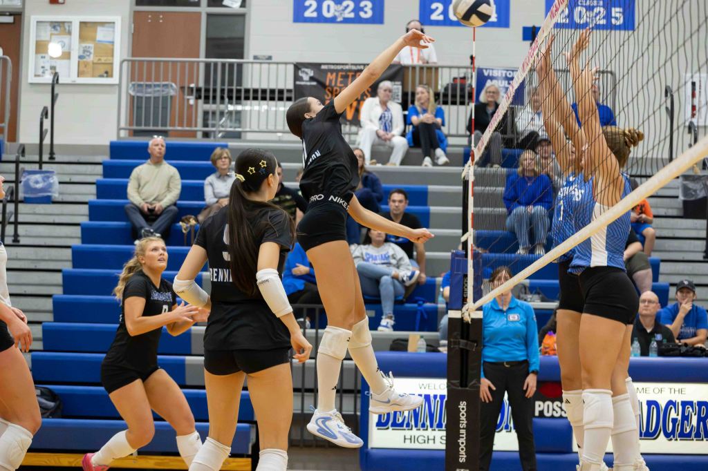 A volleyball player jumps to hit the ball over the net during a match, while teammates and opposing players are in action in the background.