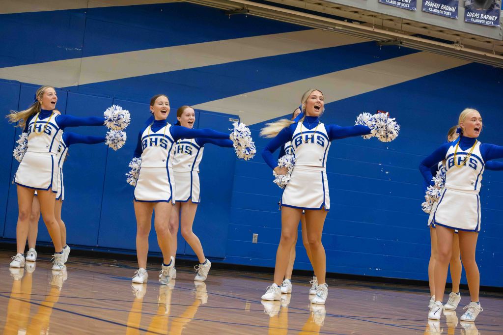 A group of cheerleaders performing in a gymnasium, wearing blue and white uniforms and holding pom-poms, with an energetic expression.