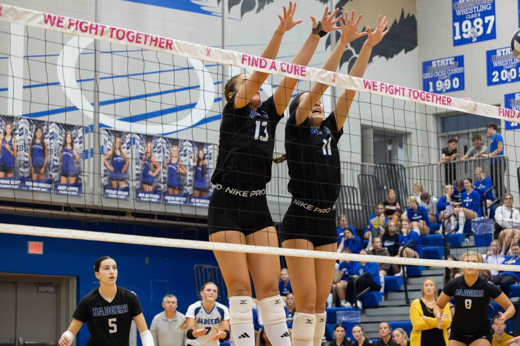 Two volleyball players block at the net during a match, while teammates and spectators are visible in the background.