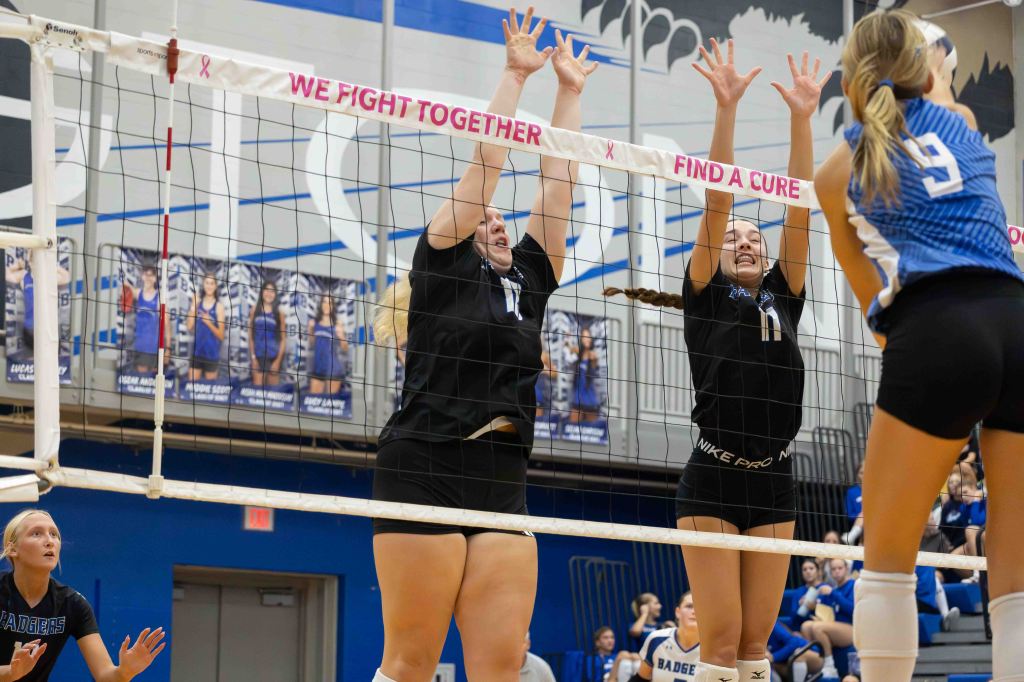 Two volleyball players in black uniforms attempting to block a spike at the net, while a player in a blue uniform prepares to hit the ball.