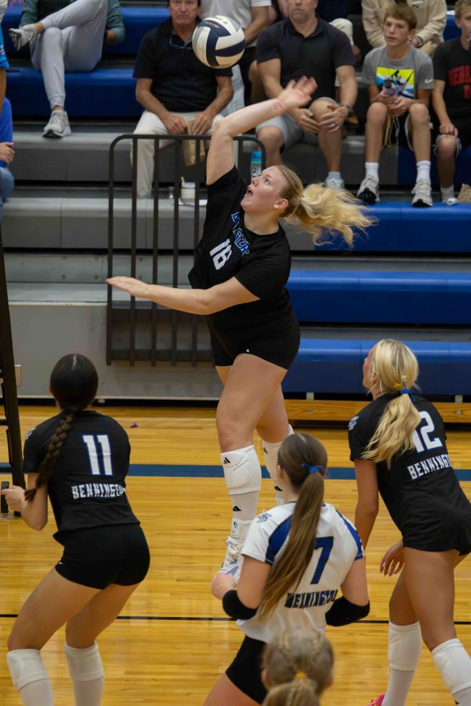 A volleyball player wearing a black jersey with the number 18 jumps to hit a volleyball, with teammates in black and white jerseys watching from below. The audience is visible in the background.