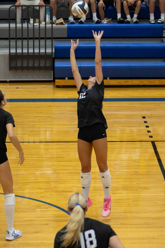 A volleyball player in a black jersey is preparing to set the ball during a game, raising both hands above her head. The background shows spectators on bleachers.
