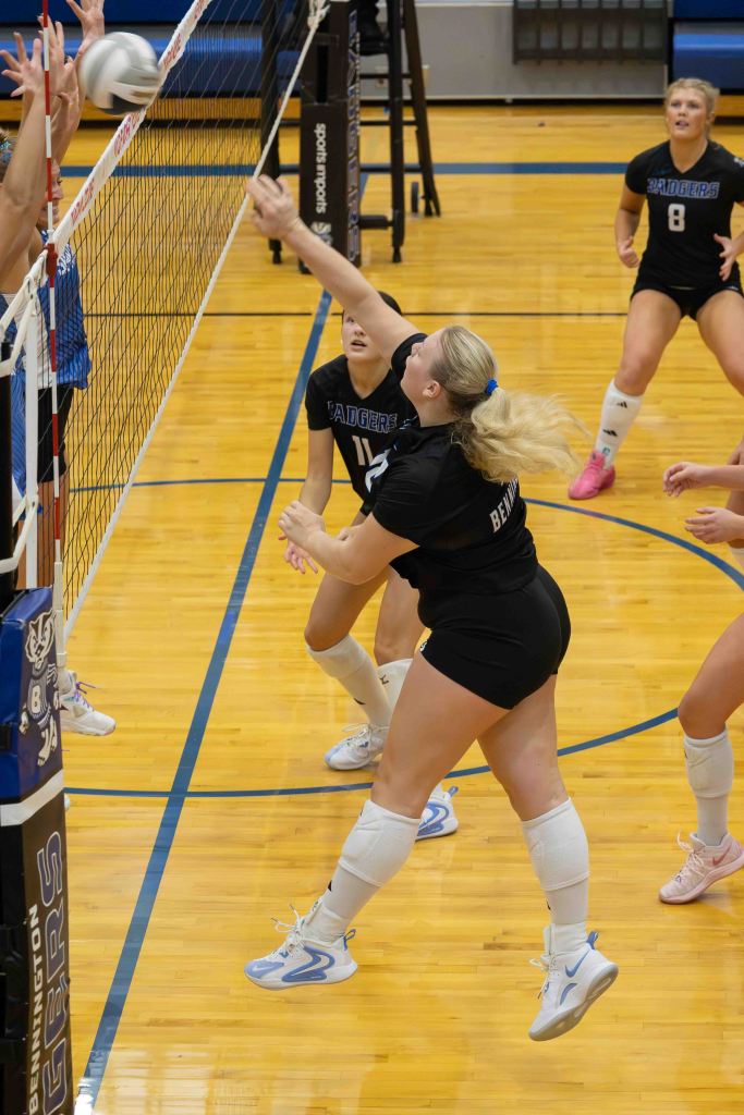 A female volleyball player in a black uniform jumps to hit the ball over the net during a match, while teammates are positioned nearby.