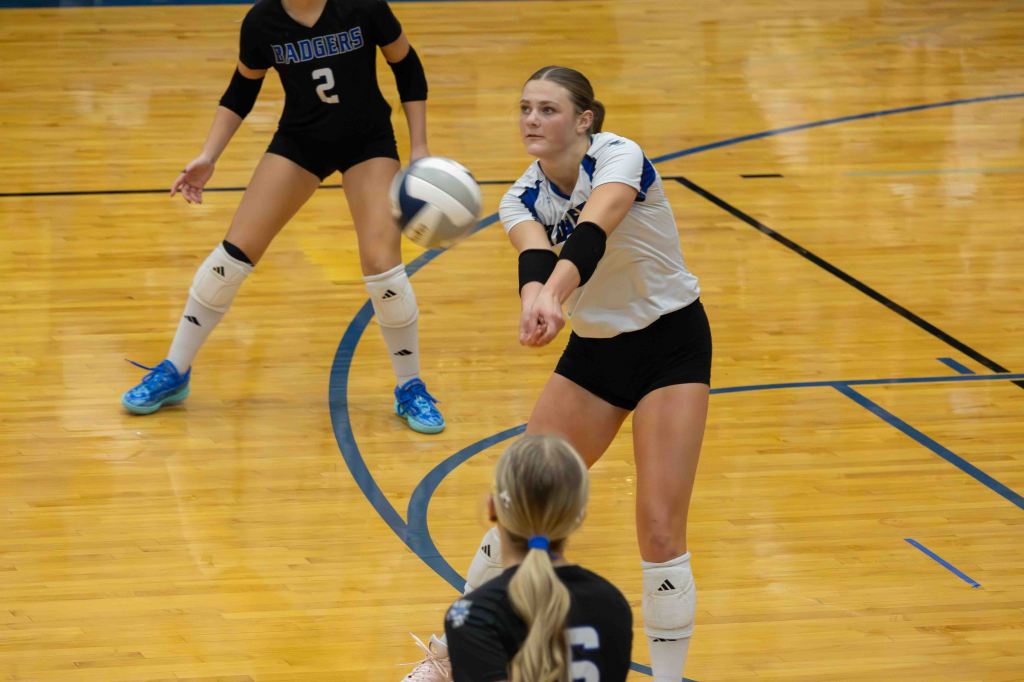 A female volleyball player prepares to receive a serve, focusing on the ball, while another teammate is positioned behind her. The court has a polished wooden floor and lines marking the boundaries.