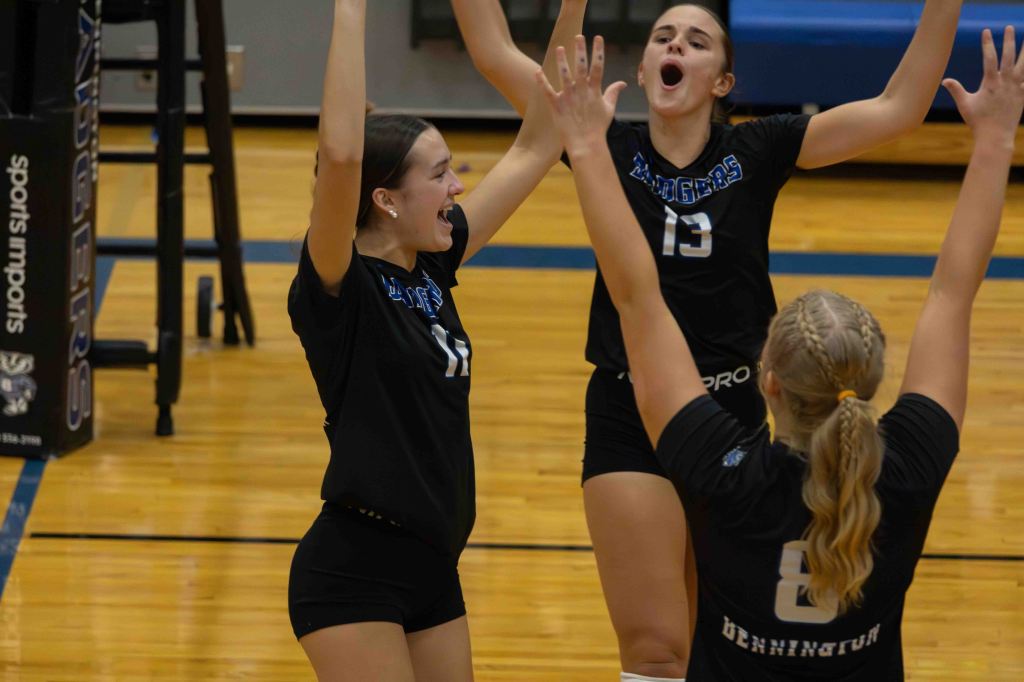 Three female volleyball players celebrating in a gym, wearing black jerseys with the number 17, 13, and 8. They are enthusiastically raising their arms and smiling.