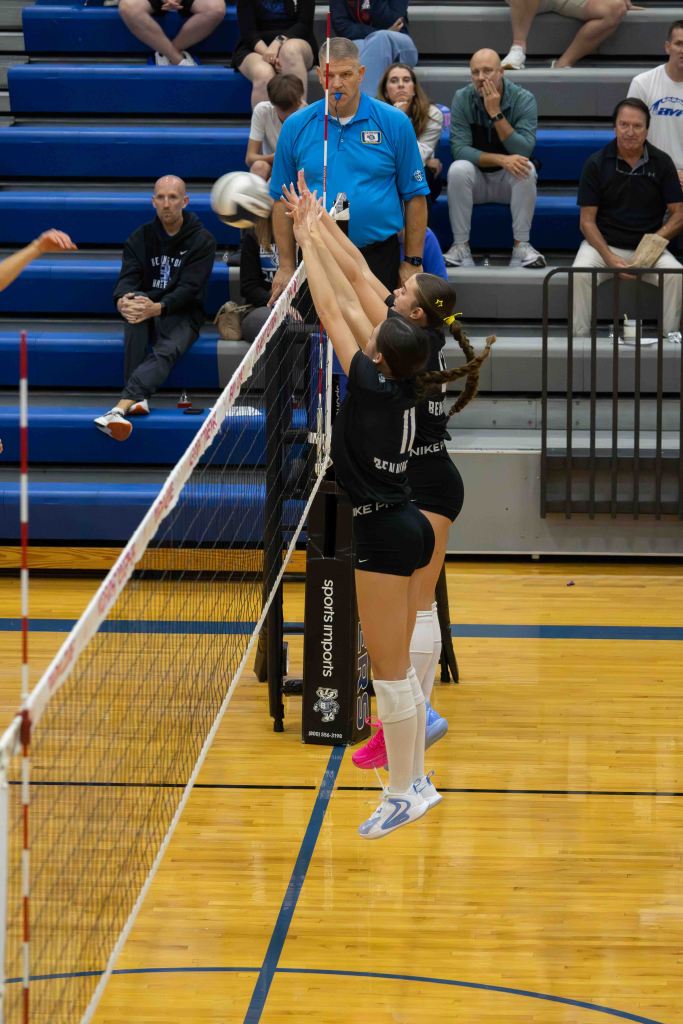 Two volleyball players jumping to block a ball at the net during a game, with an official observing and spectators in the background.