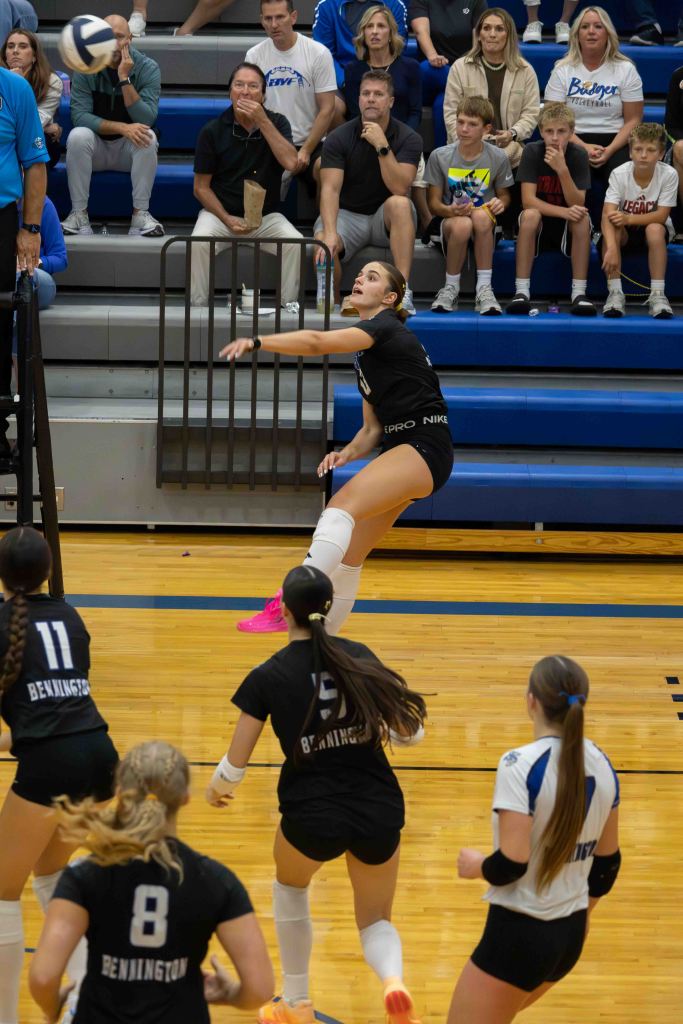 A volleyball player from the Bennington team jumps to hit the ball over the net during a game, with spectators watching in the background.