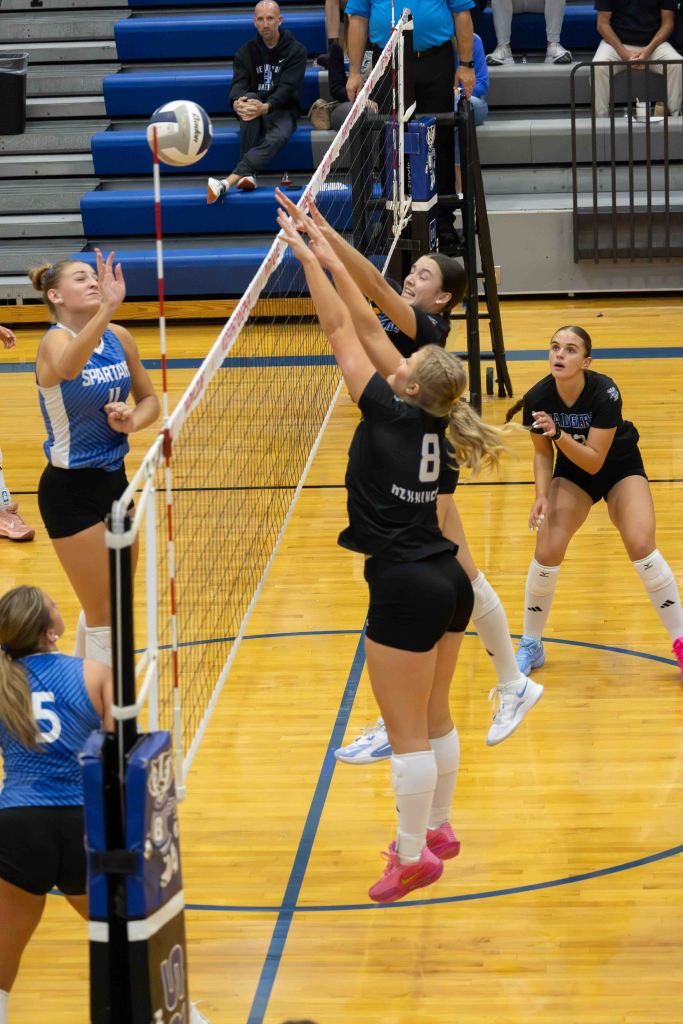 A volleyball match in progress, featuring two teams with players jumping to block a volleyball at the net. The players in black uniforms appear to be defending against an incoming hit from a player in a blue uniform.