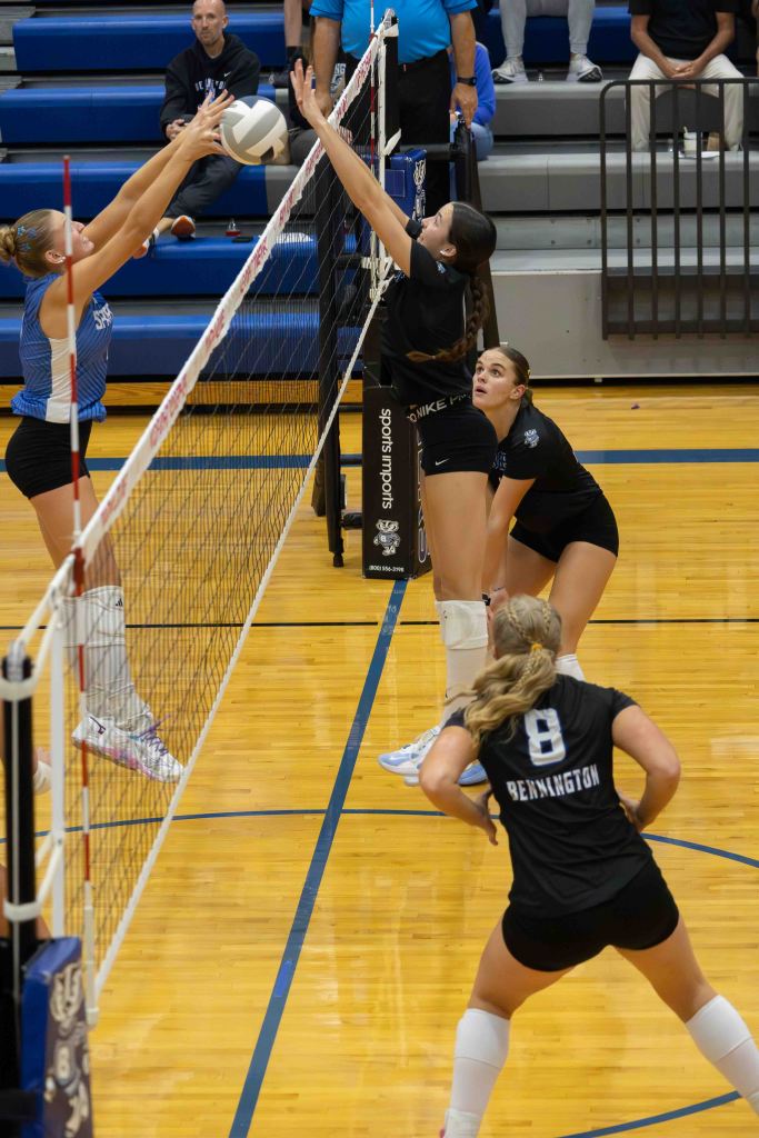 A volleyball match in action with players jumping to block a ball at the net. One player in a blue jersey is attempting to hit the ball, while two players in black jerseys are positioned to block. Another player is in a ready stance at the back.