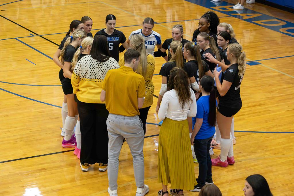 A volleyball team huddled together in a gymnasium, discussing strategies. The players are wearing matching uniforms, while a few coaches and additional individuals are also present. The wooden gym floor and bright lighting are visible in the background.