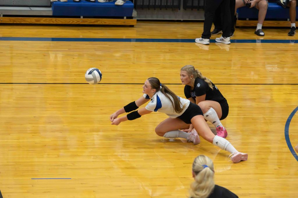 Two volleyball players on a court, one diving to play the ball while the other watches closely.