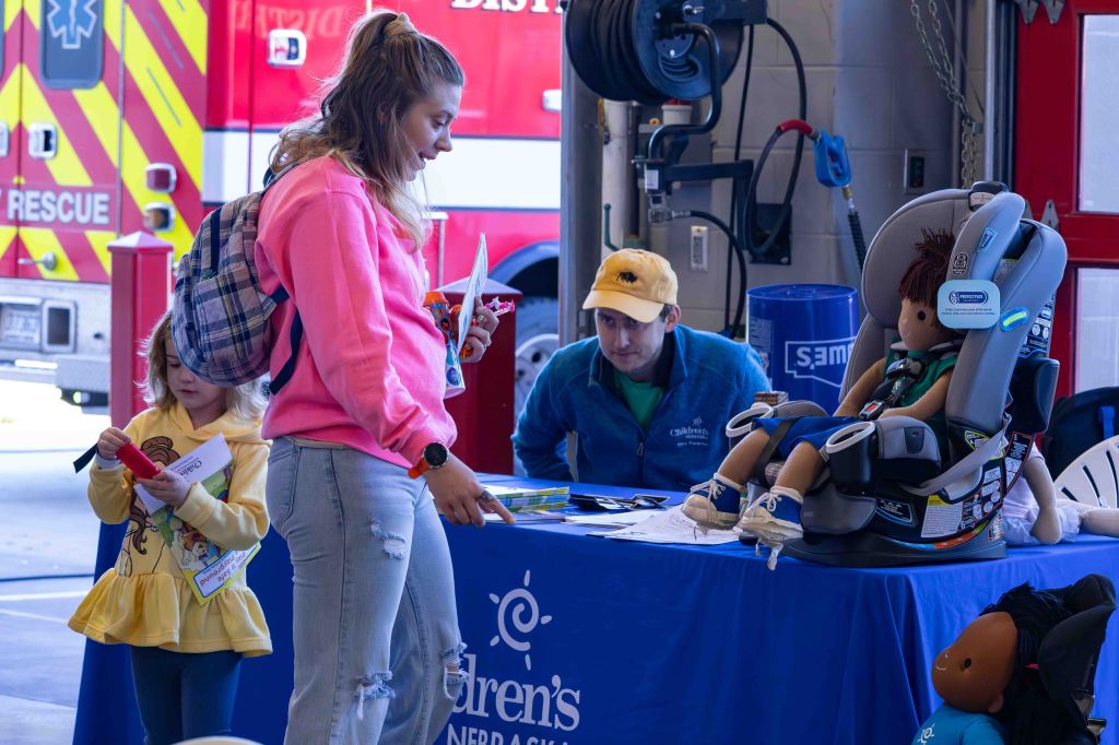 A young woman in a pink sweatshirt and a backpack examines materials at a table with children's safety information, while a young girl in a yellow sweater holds a paper. A man in a blue jacket watches attentively, and a car seat with a doll is displayed on the table. In the background, a fire truck is visible.