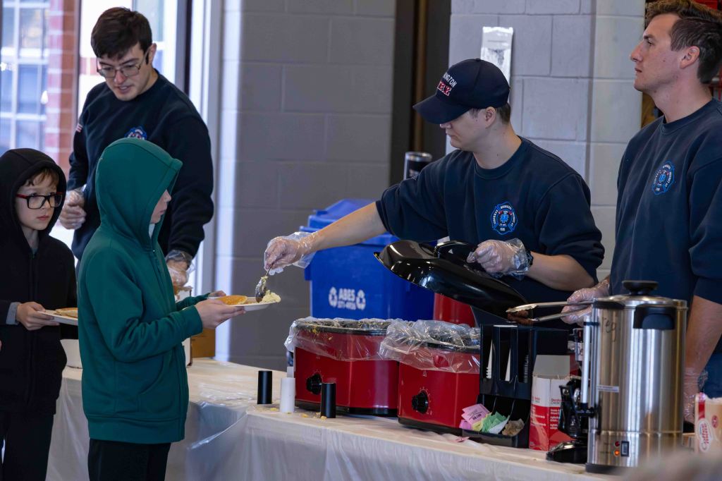 A group of children and volunteers serving food at a community event. Volunteers are wearing blue sweatshirts and gloves, while children in hoodies are holding plates as they receive meals.