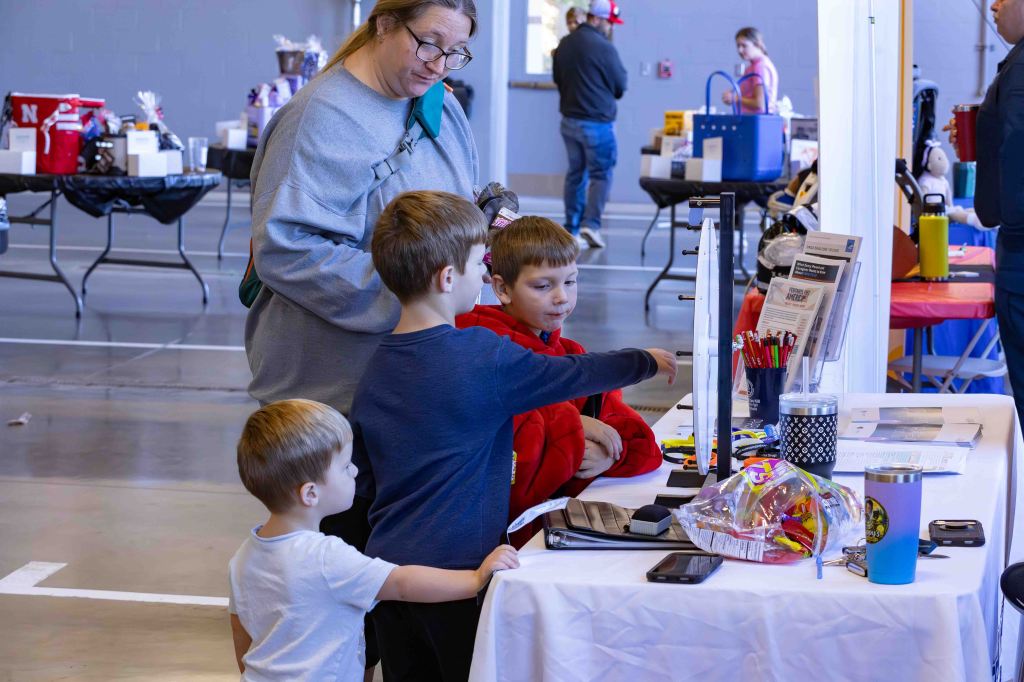 A woman and two boys are engaged at a table during an event. The woman supervises while one boy points at items on the table, which include pens, paper, and a bowl of candy. There are other tables and attendees visible in the background.