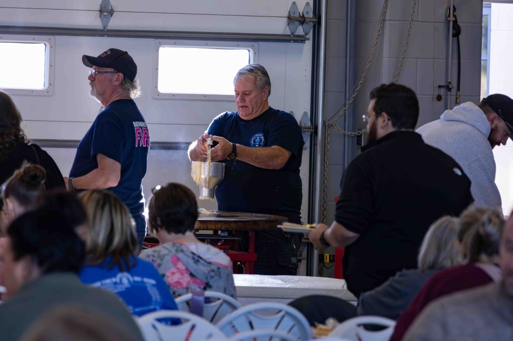 Volunteers preparing food in a community kitchen, with several people seated at tables in the background.