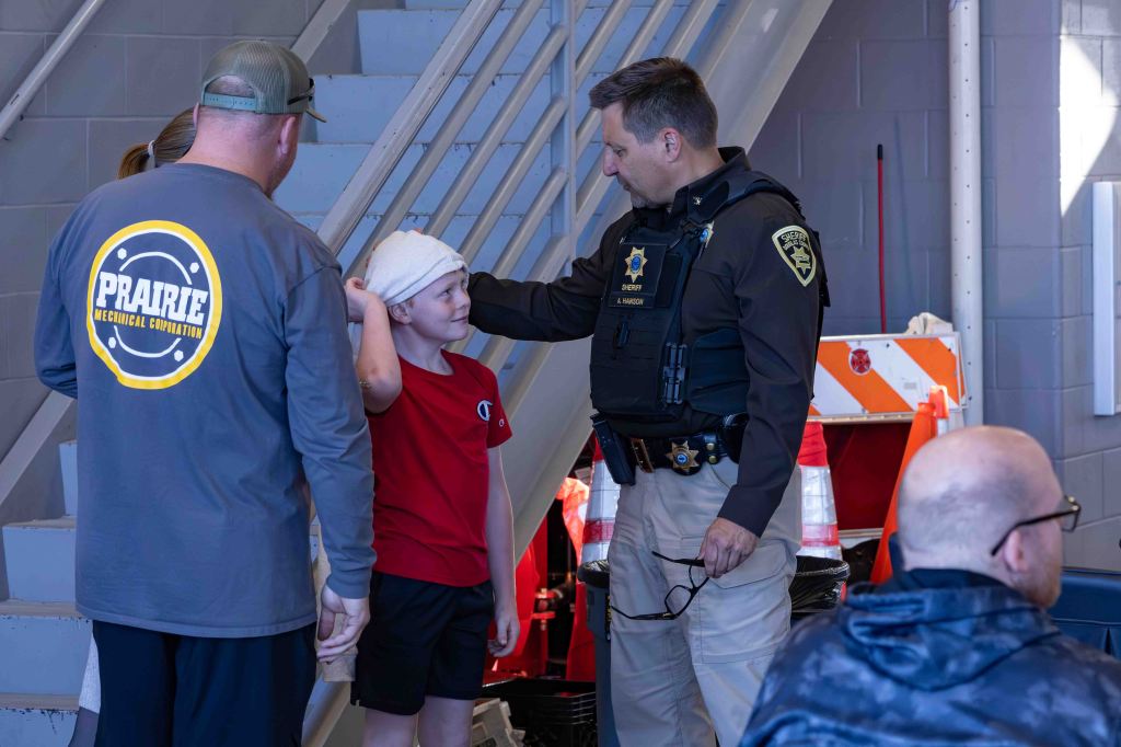 A police officer in uniform speaks to a young boy with a bandaged head, while an adult man stands nearby, both looking engaged in the conversation. In the background, there are construction cones and a staircase.