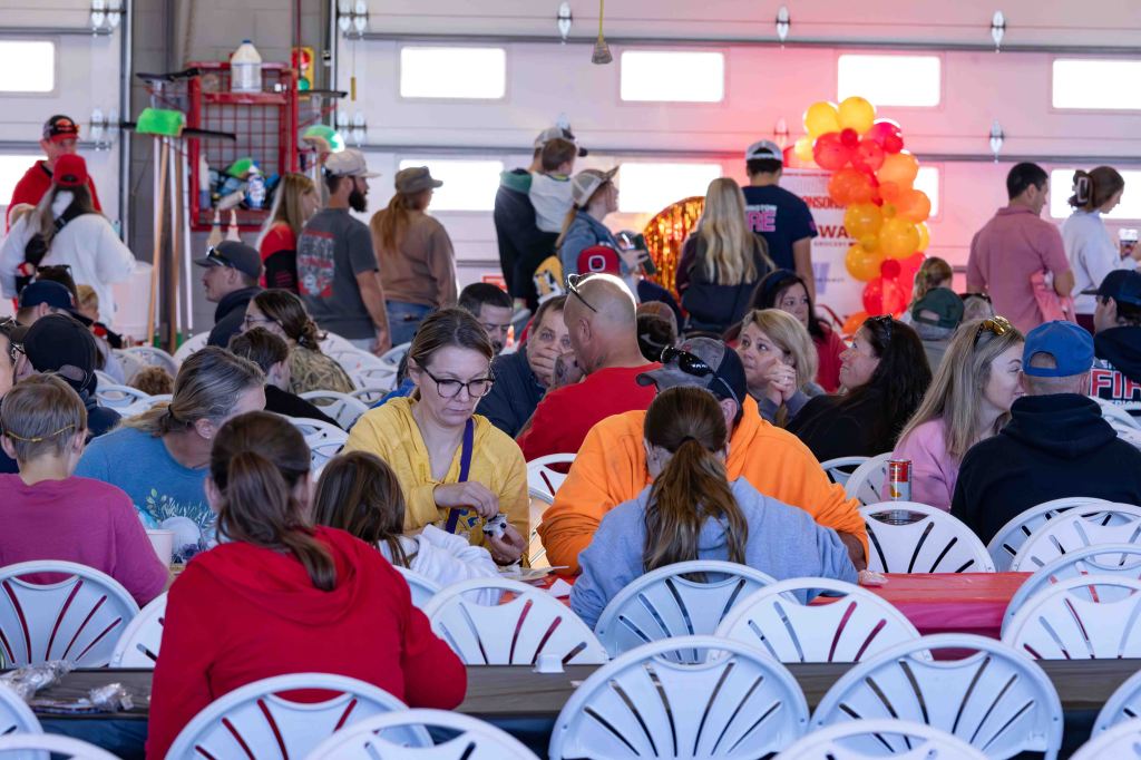 A crowded indoor event with people sitting at long tables, eating and socializing, while others stand behind them. A colorful balloon arch is visible in the background.
