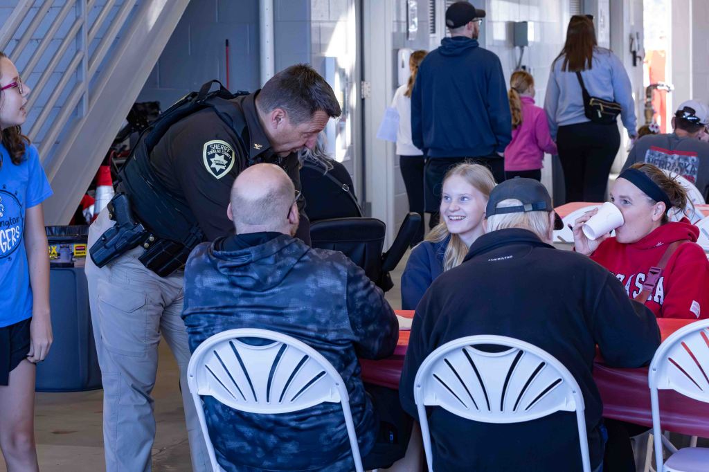 A sheriff's deputy engages with a group of people sitting at a table during a community event, as others interact in the background.