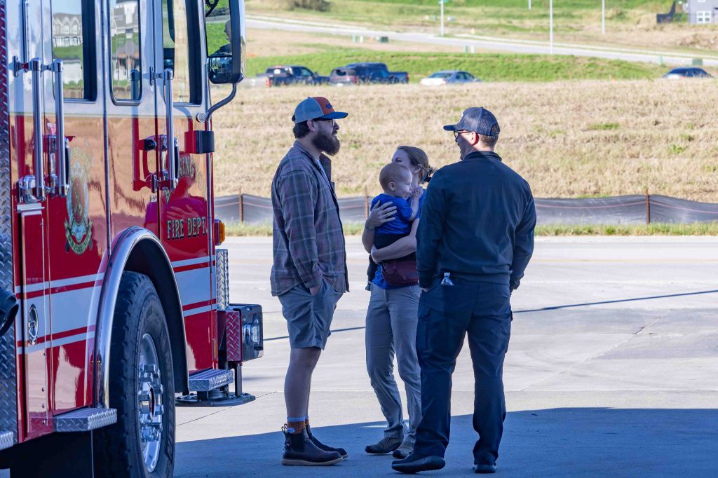 A fire truck in the foreground with a group of four people talking. One person is holding a baby, while the others engage in conversation. The background features a grassy area and parked cars.