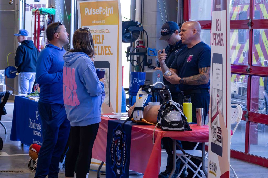 A group of people engaging at a public safety event, discussing emergency response and CPR training while standing near informational booths and equipment.