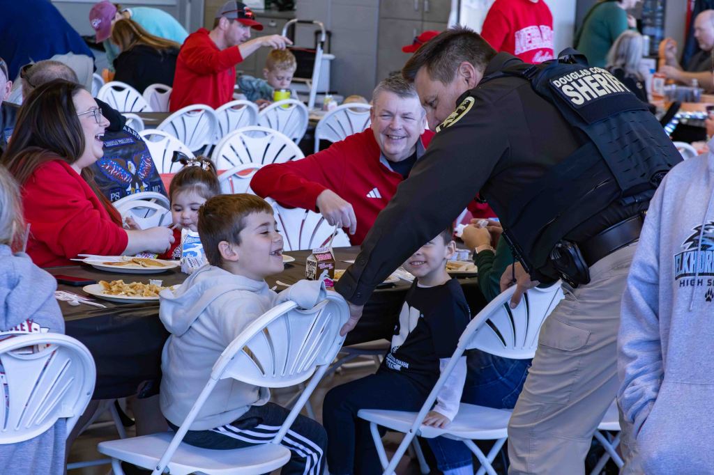 A sheriff interacts with children at a community gathering, while families share a meal at tables. Everyone appears engaged and happy.