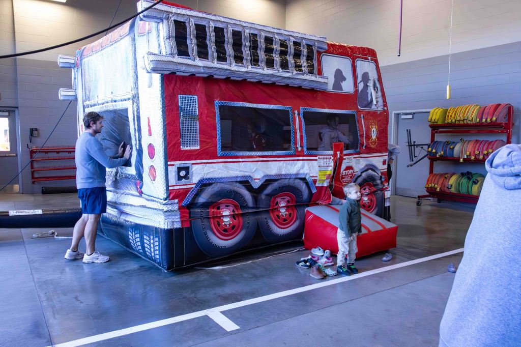 A man and a child interacting with an inflatable fire truck in a fire station, with fire hoses and other equipment visible in the background.