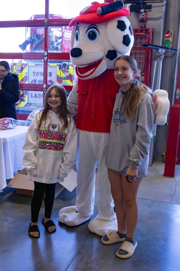 Two young girls posing with a costumed character resembling a dog in a red fireman outfit at a fire station.
