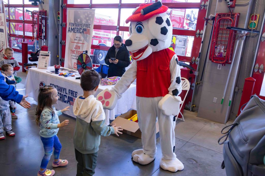 Children interacting with a costumed character resembling a fire dog at a community event, with a table promoting fentanyl awareness in the background.