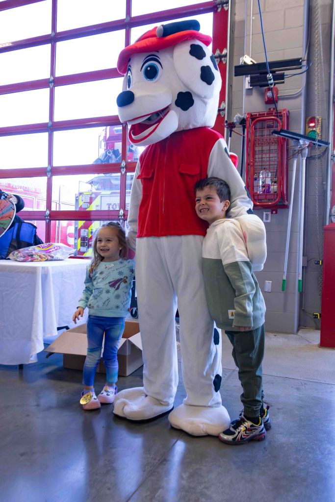 Two children posing with a character in a large dog costume inside a fire station, with red garage doors in the background.