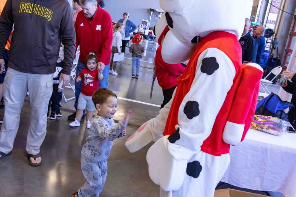 A young child in pajamas smiles and waves at a large cartoon dog mascot dressed in white with black spots, while adults and other children look on in a lively indoor setting.