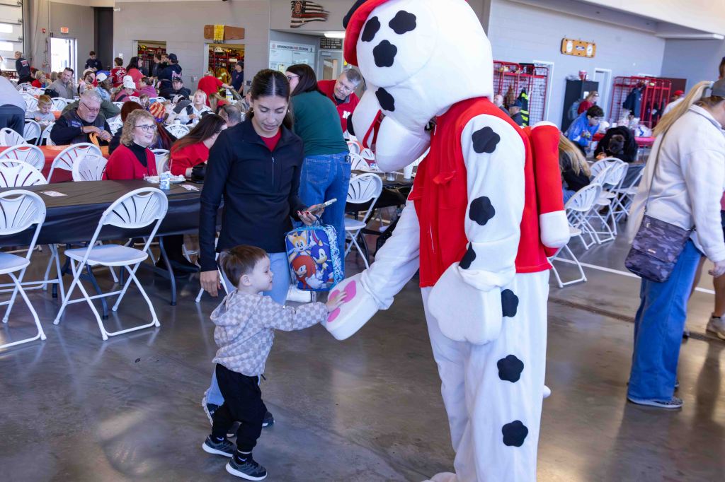 A child in a gray sweater interacts with a costumed character resembling a dalmatian at a festive indoor event. An adult woman stands beside the child, smiling.