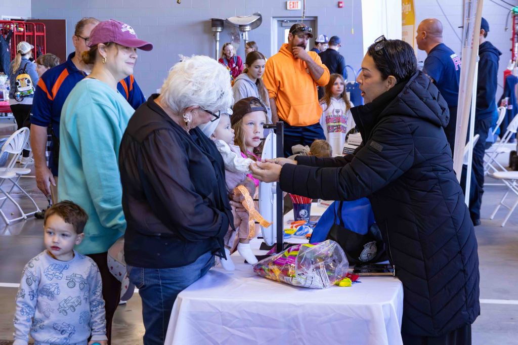 A woman in a black coat hands an item to an older woman with white hair and glasses, while a child and other people observe in a busy indoor event space.