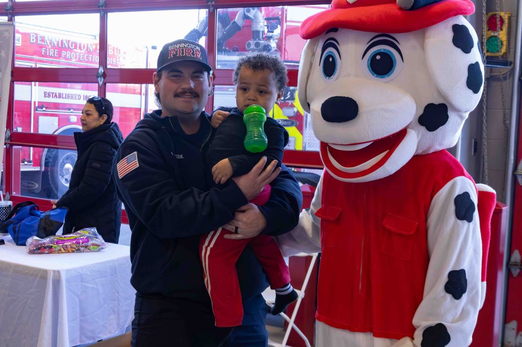 A firefighter holding a young child with a green drink, standing next to a costumed character resembling a dog. A woman is seen in the background, along with fire trucks visible through the garage doors.