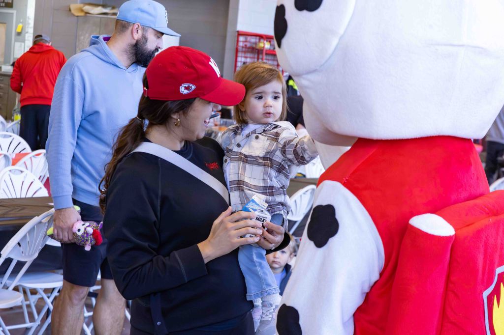 A woman holding a young child engages with a cartoonish dog mascot at a lively event. The child appears curious and is looking at the mascot, while other attendees are gathered in the background.