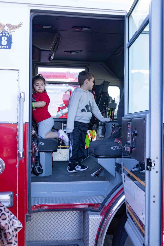Two children inside a fire truck, one sitting on a seat and the other standing near the controls.