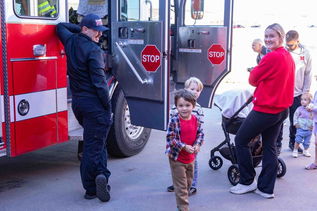 A group of children and adults gathered around a fire truck, with a firefighter standing nearby. The children are smiling and interacting, while adults chat and observe. The fire truck features visible 'STOP' signs.