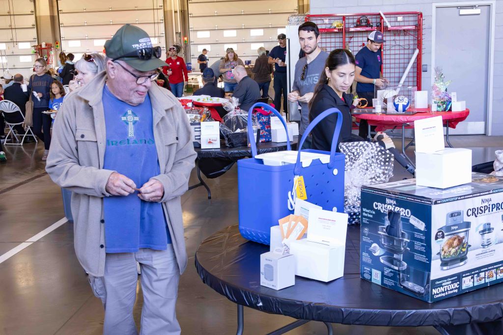 A group of people gathered at a community event with tables displaying various items for a raffle or prize giveaways. An older man in a cap and glasses stands near a blue cooler, while a woman examines items on a table with a mix of gifts including kitchen appliances.