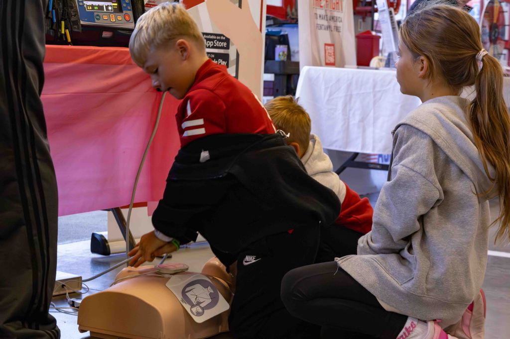 Two children are practicing CPR on a training manikin at an event, while a third child observes nearby.
