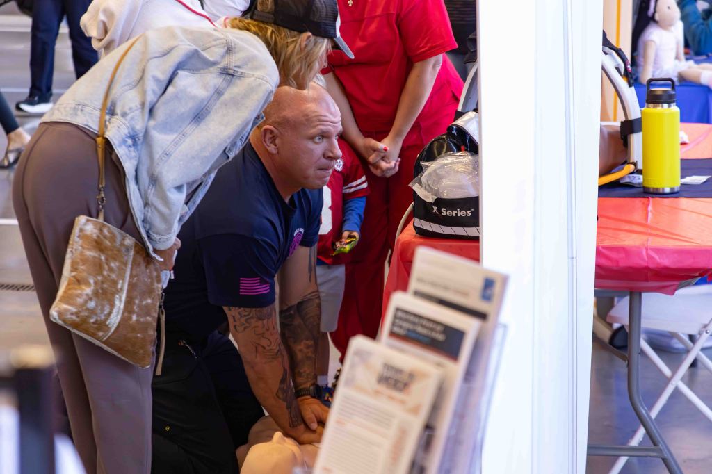 A man performing CPR on a training dummy while a group of onlookers, including a woman in a grey sweatshirt and a person in red scrubs, observe at an educational event.