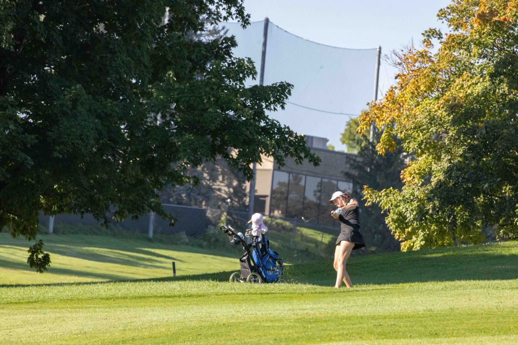 A female golfer swings a club on a grassy course, with a golf bag nearby and trees in the foreground.