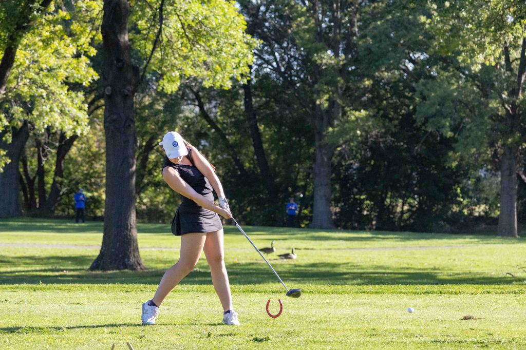 A female golfer in a black outfit swings a golf club on a green course with trees in the background.