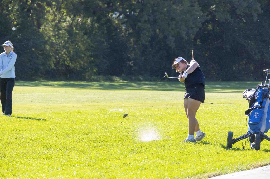 A golfer swings a club, striking the ball from the grass, with another player observing in the background.